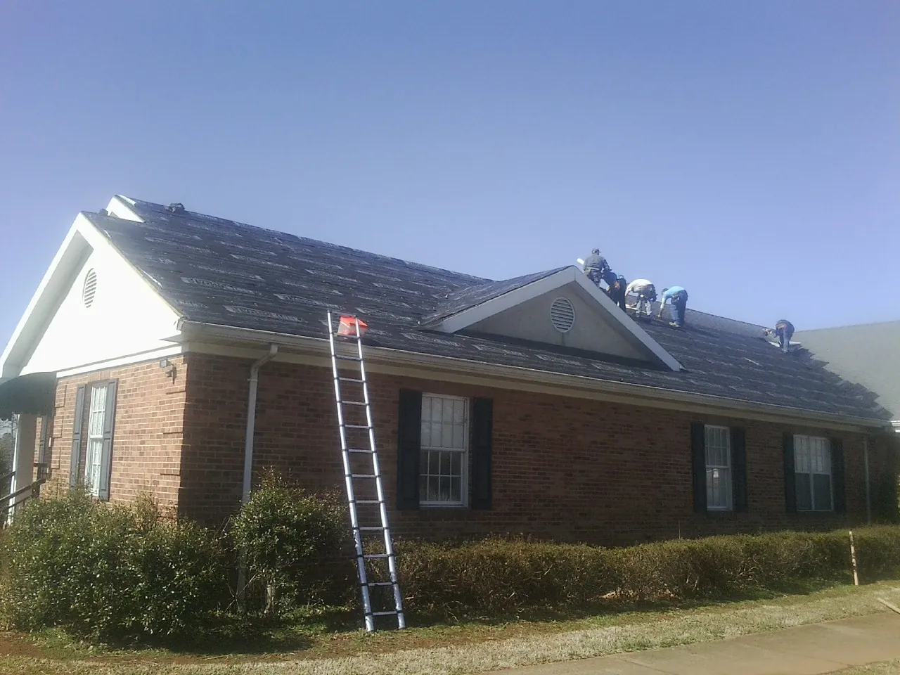 Crew tearing shingles off a brick ranch roof with a ladder set — Emergency Roof Repair work by our crew in Deferiet