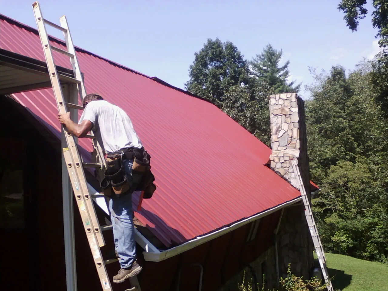 Roofer on a ladder beside a red metal roof and stone chimney — Soffit & Fascia Repair by Roofing Brothers in Deferiet
