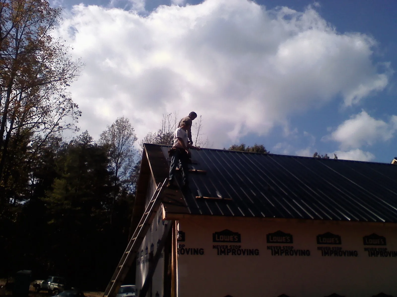 Roofer fastening dark metal panels high against a cloudy sky, from a Roof Inspection job in Deferiet